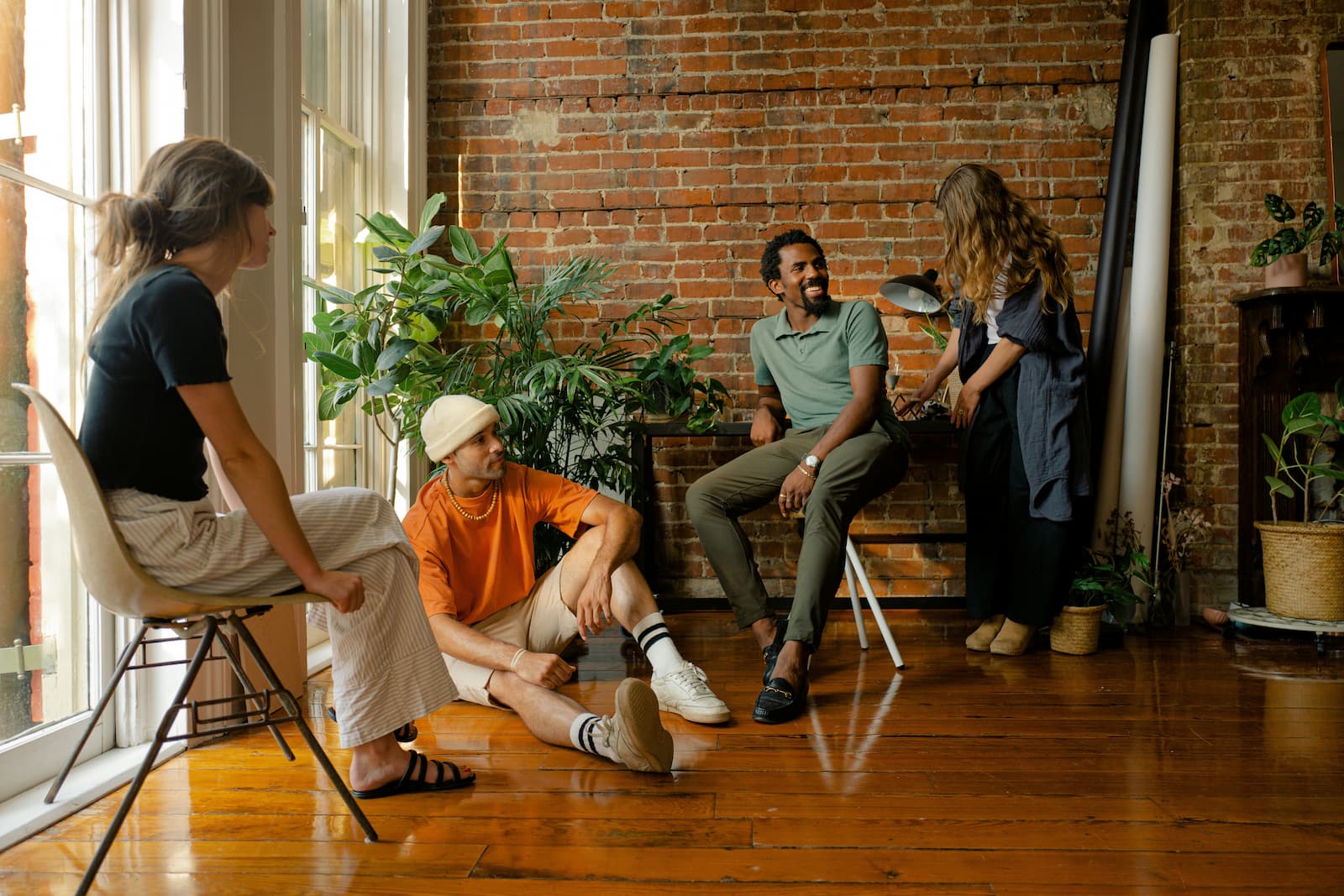 Four friends gathered in a warmly lit loft living space with exposed brick walls and large windows, talking and laughing — the kind of home that draws people in and stays with them long after they leave.