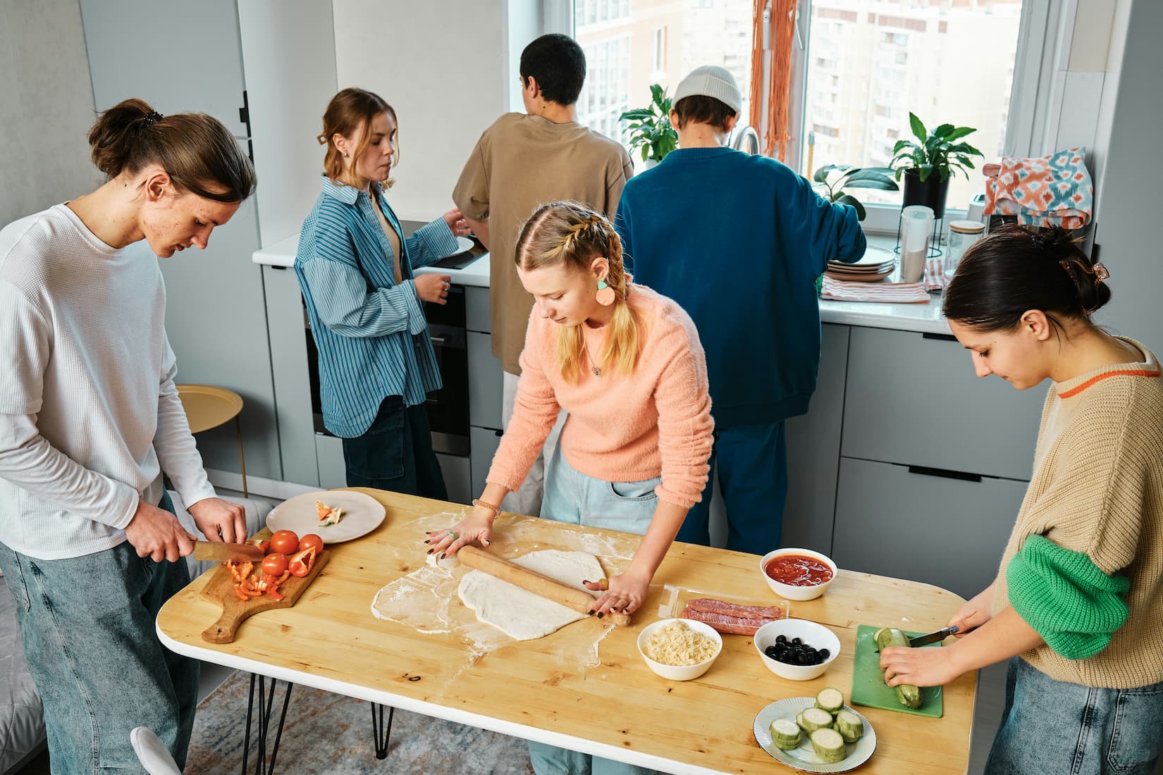 A group of young roommates cook together in a shared kitchen, rolling dough and prepping toppings, the kind of scene where a kitchen odor eliminator and knowing how to block smell from kitchen cooking keep a space guest-ready.