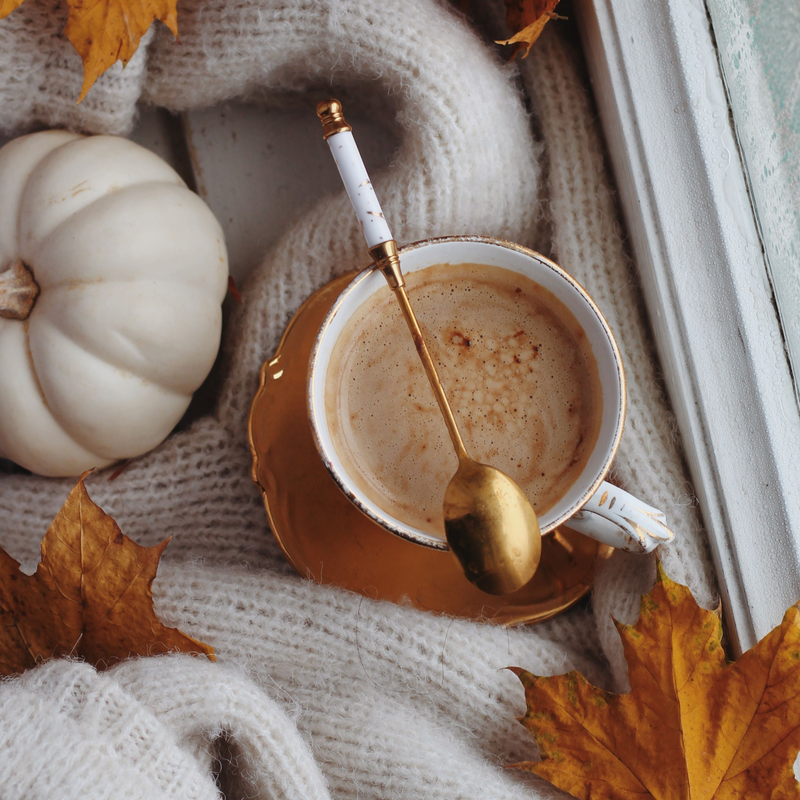 A top-down cozy autumn scene featuring a white ceramic coffee cup with a gold rim, filled with a frothy latte, resting on a matching saucer with a gold spoon. The cup is styled on a cream knit blanket next to a small white pumpkin and scattered golden maple leaves.